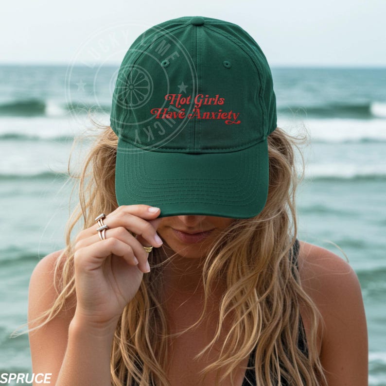 May include: A dark green baseball cap with the text "Hot Girls Have Anxiety" in red script. The cap is worn by a person with long blonde hair, with the ocean in the background.
