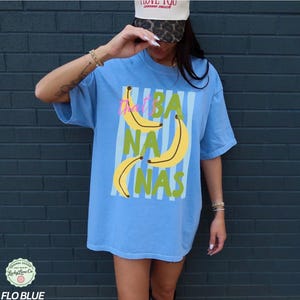 May include: A light blue t-shirt with the text "That's Bananas" in a vertical striped design. The shirt features yellow banana illustrations and green text. The shirt is a Flo Blue color.