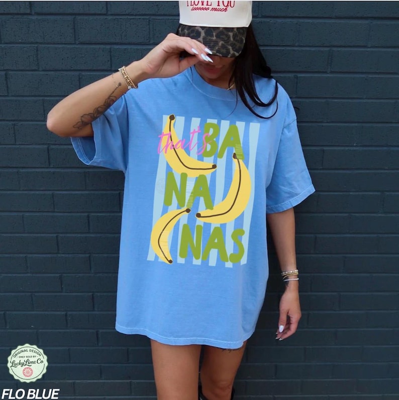 May include: A light blue t-shirt with the text "That's Bananas" in a vertical striped design. The shirt features yellow banana illustrations and green text. The shirt is a Flo Blue color.
