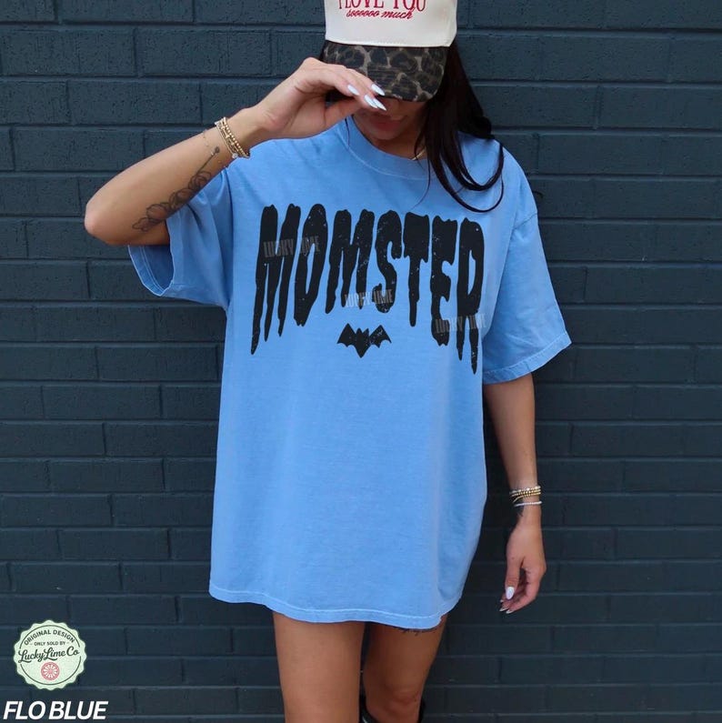 May include: A light blue t-shirt with the word "MOMSTER" in black, stylized text and a bat graphic. The shirt is a relaxed fit and has short sleeves. The text "FLO BLUE" is at the bottom of the image.