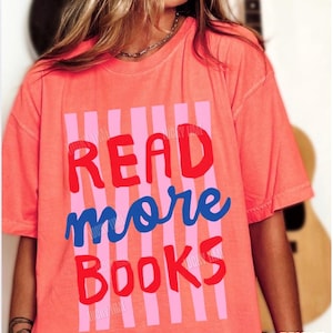 Puede incluir: Camiseta de color salm&oacute;n brillante con las palabras "READ more BOOKS" en letras rojas y azules. El texto est&aacute; sobre un fondo de rayas verticales rosas. La camiseta tiene mangas cortas.