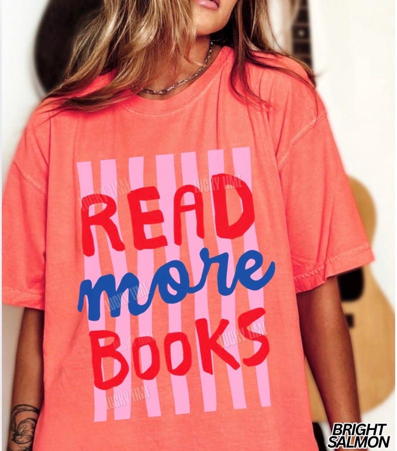 Puede incluir: Camiseta de color salm&oacute;n brillante con las palabras "READ more BOOKS" en letras rojas y azules. El texto est&aacute; sobre un fondo de rayas verticales rosas. La camiseta tiene mangas cortas.