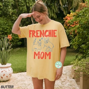 May include: A butter-colored t-shirt with the words "FRENCHIE MOM" in red, featuring blue illustrations of French Bulldogs. The shirt has a relaxed fit and a round neckline. The text "Life is better with French Bulldogs" is also on the shirt.