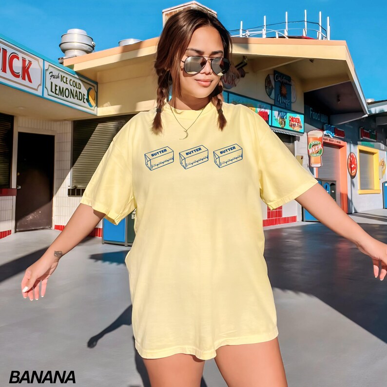Puede incluir: Una camiseta amarilla de manga corta con un gr&aacute;fico de tres barras de mantequilla. El texto "BUTTER" est&aacute; impreso en cada barra.