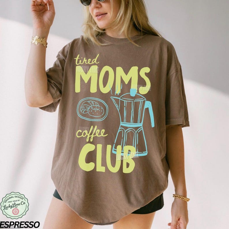 May include: Brown t-shirt with the text "tired MOMS coffee CLUB" in yellow and light blue. The design includes a coffee pot and a pastry. The shirt is a relaxed fit.