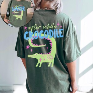 May include: Moss green t-shirt with a graphic of a green crocodile and the text "after while CROCODILE" on the back. The front of the shirt has a smaller graphic with the text "later GATOR". The design includes pink and blue accents.