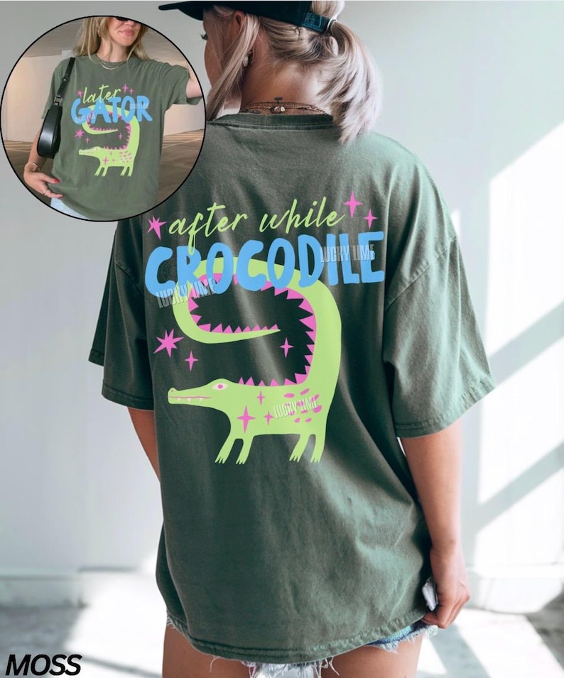 May include: Moss green t-shirt with a graphic of a green crocodile and the text "after while CROCODILE" on the back. The front of the shirt has a smaller graphic with the text "later GATOR". The design includes pink and blue accents.