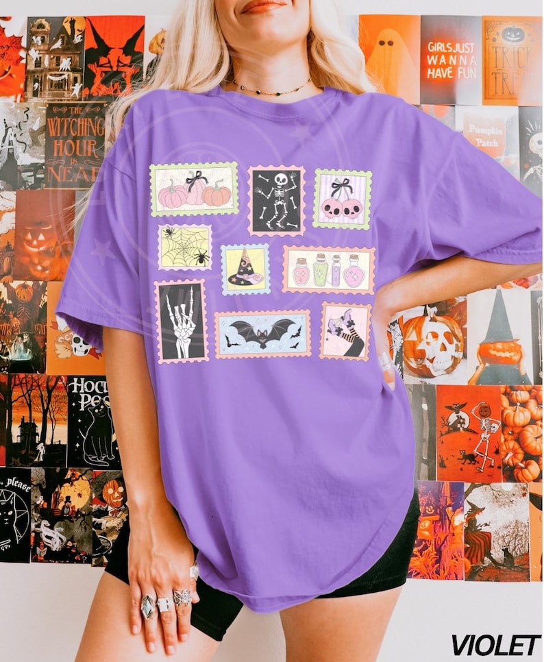 May include: A violet-colored t-shirt featuring a collection of Halloween-themed postage stamp designs. The stamps depict pumpkins, skeletons, bats, and other spooky elements. The word "VIOLET" is printed at the bottom right.