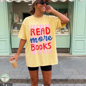 Puede incluir: Camiseta amarilla con el texto "READ more BOOKS" en rojo, azul y rosa. La camiseta se lleva con pantalones cortos negros. La imagen est&aacute; tomada al aire libre.
