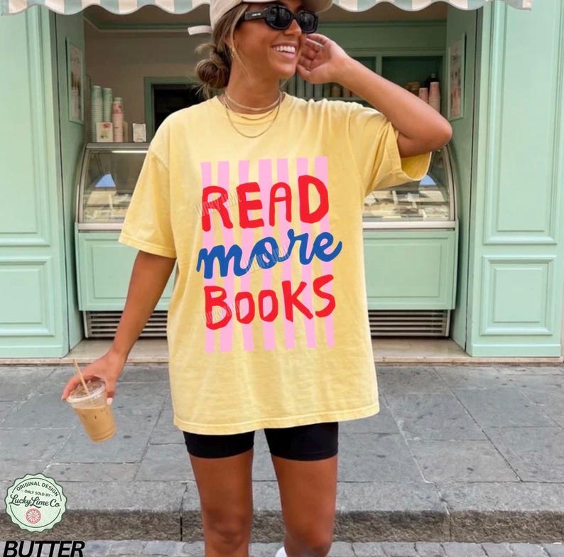 Puede incluir: Camiseta amarilla con el texto "READ more BOOKS" en rojo, azul y rosa. La camiseta se lleva con pantalones cortos negros. La imagen est&aacute; tomada al aire libre.