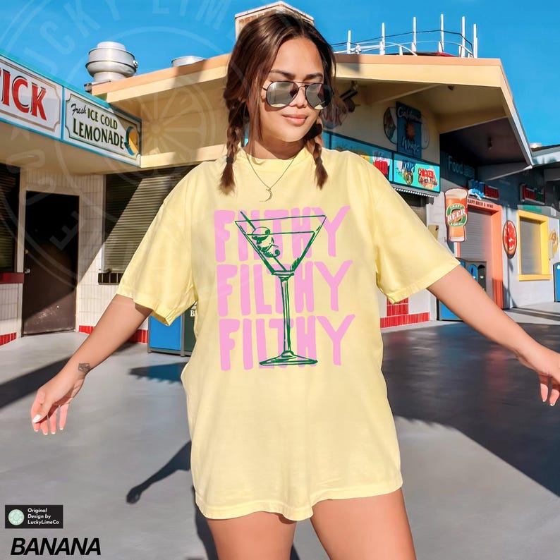 Pode incluir: Uma camiseta amarela de manga curta com um gr&aacute;fico rosa de uma ta&ccedil;a de martini com azeitonas e o texto "Filthy" repetido tr&ecirc;s vezes.