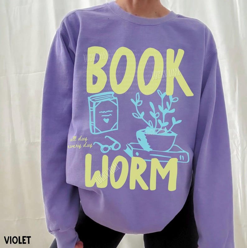 May include: Violet sweatshirt with the words "BOOK WORM" in yellow. The design includes a book, glasses, and a potted plant. The text "all day every day" is also printed on the sweatshirt.