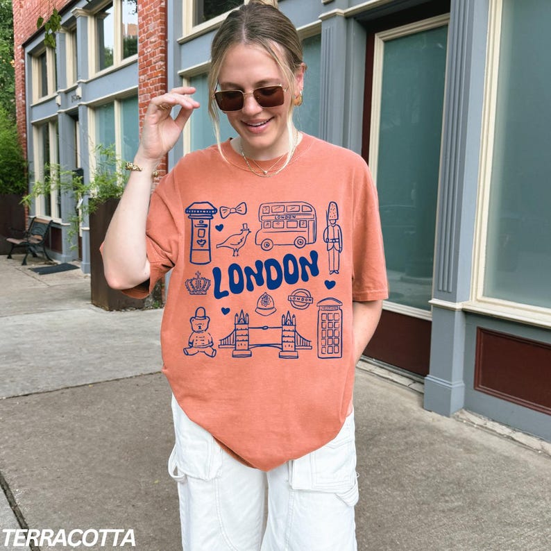May include: A terracotta orange t-shirt with a blue graphic design of London landmarks, including a double-decker bus, a red telephone booth, Big Ben, and the Tower Bridge. The text "LONDON" is printed in blue on the shirt.