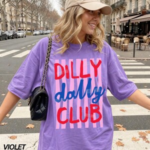 May include: A violet-colored t-shirt with the words "DILLY DALLY CLUB" in red and blue lettering, set against a pink striped background. The shirt is worn with a beige cap and a black quilted shoulder bag.