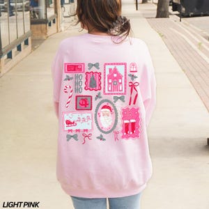 May include: Light pink sweatshirt with a festive Christmas-themed design on the back. The design includes various pink and gray postage stamp-style images with holiday elements like Santa Claus, a gingerbread house, and candy canes.