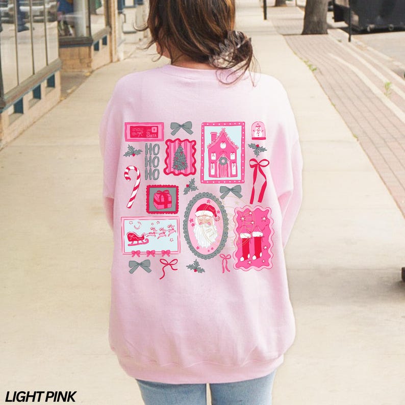 May include: Light pink sweatshirt with a festive Christmas-themed design on the back. The design includes various pink and gray postage stamp-style images with holiday elements like Santa Claus, a gingerbread house, and candy canes.