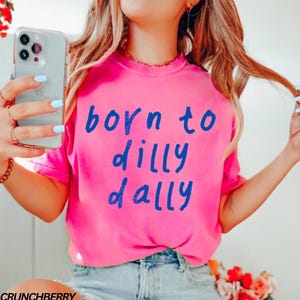 May include: A bright pink t-shirt with the words "born to dilly dally" in blue lettering. The shirt is made of a soft material and is a casual style. The shirt is a vibrant color and is perfect for everyday wear.