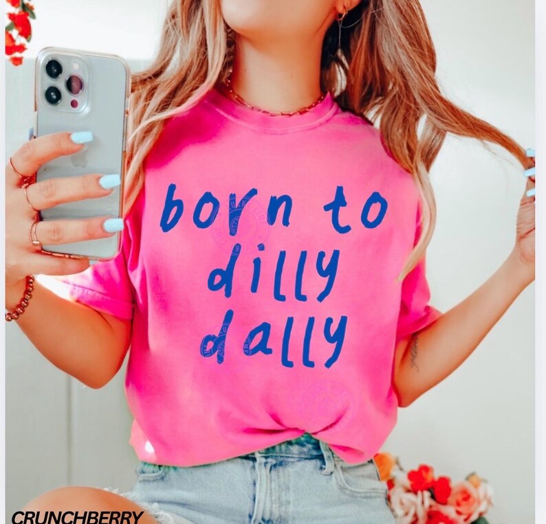 May include: A bright pink t-shirt with the words "born to dilly dally" in blue lettering. The shirt is made of a soft material and is a casual style. The shirt is a vibrant color and is perfect for everyday wear.