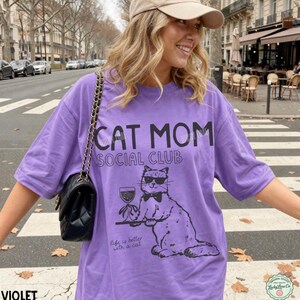 K&ouml;nnte beinhalten: Ein violettes kurz&auml;rmeliges T-Shirt mit einem schwarzen Grafikdruck einer Katze, die eine Sonnenbrille und eine Fliege tr&auml;gt und ein Weinglas auf einem Tablett h&auml;lt. Der Text lautet CAT MOM SOCIAL CLUB und Life is better with a cat. Eine schwarze Tasche ist zu sehen.