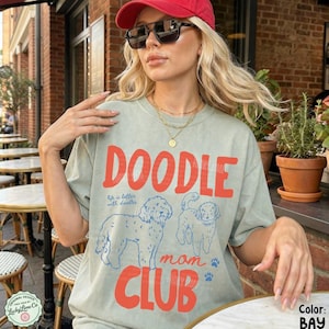 Puede incluir: Camiseta verde claro con las palabras "DOODLE mom CLUB" en rojo. La camiseta presenta dos ilustraciones de perros doodle en azul y la frase "life is better with doodles". La camiseta es de corte holgado.