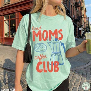 May include: A light mint green t-shirt featuring a graphic print in red and blue. The text reads tired MOMS coffee CLUB. The design includes a line drawing of a plate with a pastry and a moka pot coffee maker. A black shoulder bag is worn.