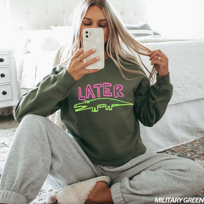 May include: An olive green sweatshirt featuring the word "LATER" in pink and a neon green alligator design. The sweatshirt is worn with grey sweatpants. The image is taken indoors.