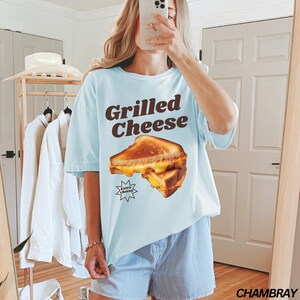 May include: A light blue t-shirt with a graphic of a grilled cheese sandwich and the text "Grilled Cheese" in brown letters. The text "Extra Cheesy" is in a smaller font below the sandwich. The shirt is being worn by a person with long brown hair.