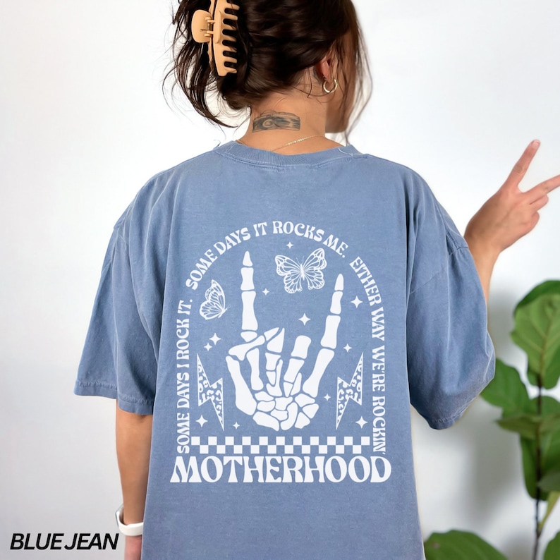 May include: Blue jean-colored t-shirt with a white graphic on the back. The graphic includes a skeleton hand in a rock-and-roll gesture, butterflies, lightning bolts, and the word "MOTHERHOOD". Text reads "SOME DAYS I ROCK IT. SOME DAYS IT ROCKS ME. EITHER WAY WE'RE ROCKIN".