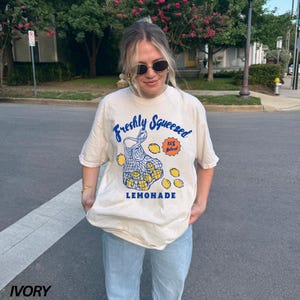 Puede incluir: Una mujer lleva una camiseta beige claro con un gr&aacute;fico de limones en una bolsa de malla y el texto "Freshly Squeezed 100% Natural LEMONADE".