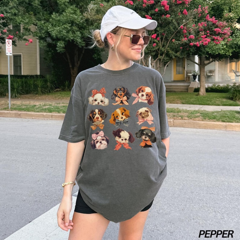 May include: A dark gray oversized t-shirt with a graphic print of 12 different dogs wearing bows. The dogs are all different breeds and have different expressions. The shirt is being worn by a person with a white baseball cap and sunglasses.
