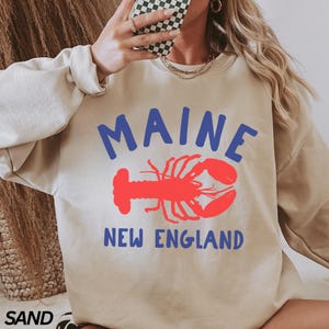 May include: A beige sweatshirt with a blue and red graphic of a lobster and the text "MAINE NEW ENGLAND".