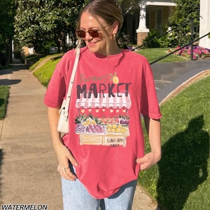 May include: A red t-shirt with a "Farmer's Market" graphic featuring a fruit stand with various fruits and the text "10AM-5PM DAILY". The shirt is a watermelon color.