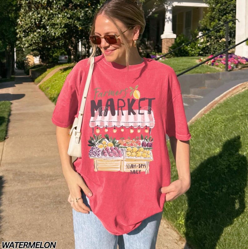 May include: A red t-shirt with a "Farmer's Market" graphic featuring a fruit stand with various fruits and the text "10AM-5PM DAILY". The shirt is a watermelon color.