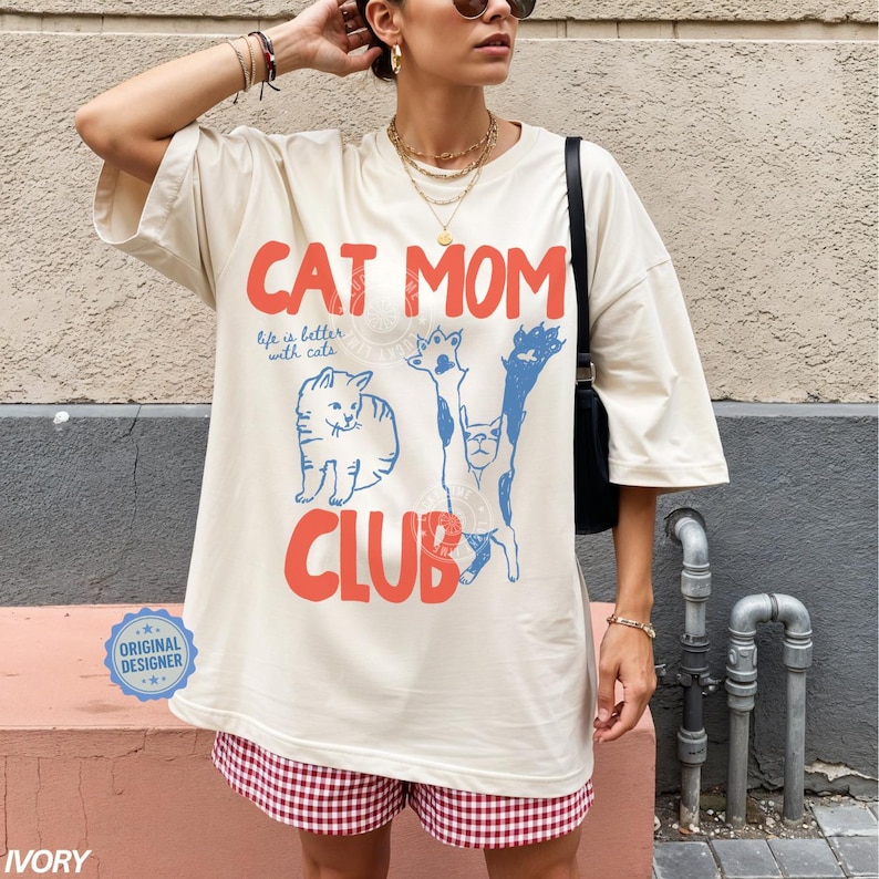 Puede incluir: Camiseta de gran tama&ntilde;o de color marfil con el texto "CAT MOM CLUB" en rojo y azul, con ilustraciones de gatos. La camiseta tambi&eacute;n tiene el texto "life is better with cats". La camiseta se combina con pantalones cortos a cuadros rojos y blancos.