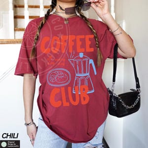 Op de afbeelding: Rood T-shirt met een afbeelding van een koffiepot en donuts. De tekst op het shirt luidt "Coffee Club" en "all day every day".