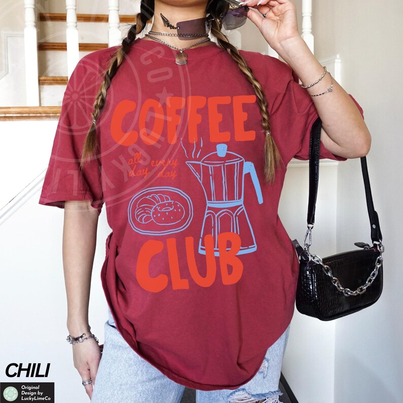 Op de afbeelding: Rood T-shirt met een afbeelding van een koffiepot en donuts. De tekst op het shirt luidt "Coffee Club" en "all day every day".