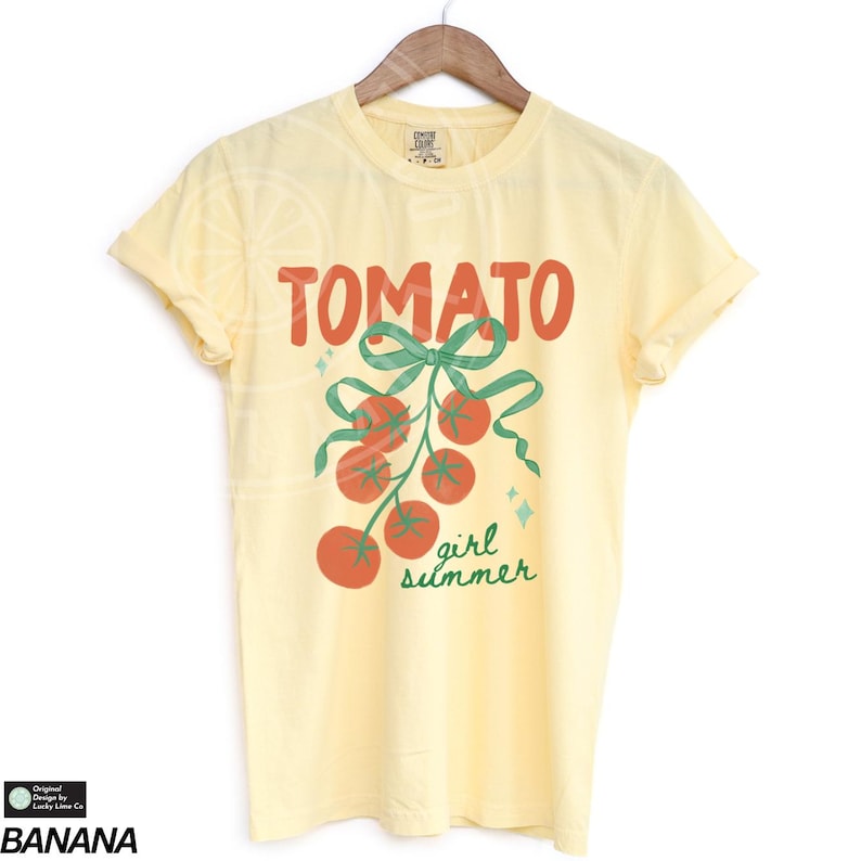 May include: A light yellow t-shirt with the word "TOMATO" in red block letters above a green bow and a cluster of red tomatoes. The words "girl summer" are in green script below the tomatoes. The shirt is hanging on a wooden hanger.