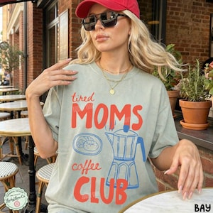 May include: A sage green t-shirt featuring the red text tired MOMS coffee CLUB. The design includes a blue line drawing of a moka pot and a plate with a pastry. The shirt has a crew neckline and short sleeves, displaying a casual graphic print.