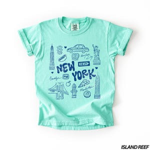 May include: A light turquoise t-shirt with a blue graphic design featuring iconic New York City landmarks and text. The design includes the words "New York", the Brooklyn Bridge, the Statue of Liberty, and other city symbols.