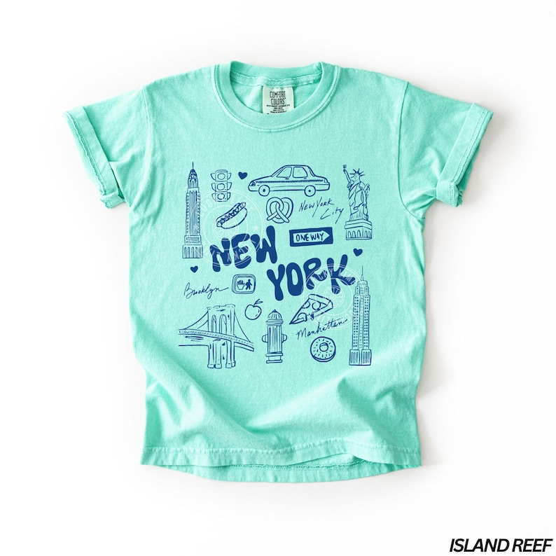 May include: A light turquoise t-shirt with a blue graphic design featuring iconic New York City landmarks and text. The design includes the words "New York", the Brooklyn Bridge, the Statue of Liberty, and other city symbols.