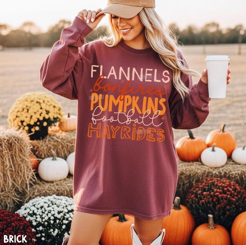 May include: A long-sleeve maroon sweatshirt featuring the text FLANNELS, bonfires, PUMPKINS, football, and HAYRIDES in a mix of block and script fonts. The garment has a crew neckline and a relaxed fit, with the text printed in orange and white.