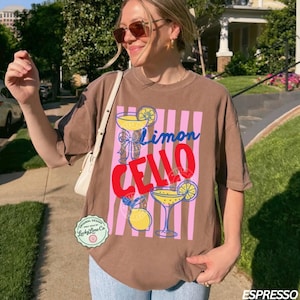 May include: Espresso-colored t-shirt with a graphic design featuring the words "Limon Cello" in blue and red, along with illustrations of cocktails and lemons. The shirt has a relaxed fit and short sleeves.