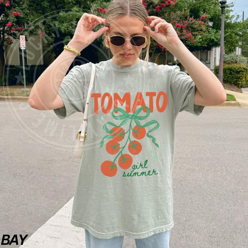 May include: A light green t-shirt with the word "TOMATO" in orange block letters. Below the word are orange tomatoes with green stems and a green bow. The words "girl summer" are below the tomatoes.