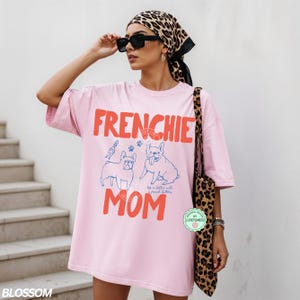 May include: Pink t-shirt with the words "FRENCHIE MOM" in red, along with blue illustrations of French Bulldogs. The shirt is part of a casual wear collection. The model is wearing sunglasses and a leopard print scarf.