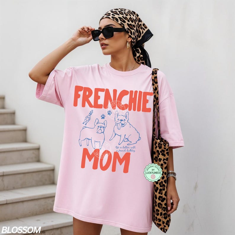 May include: Pink t-shirt with the words "FRENCHIE MOM" in red, along with blue illustrations of French Bulldogs. The shirt is part of a casual wear collection. The model is wearing sunglasses and a leopard print scarf.
