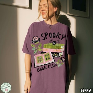 Op de afbeelding: Bessenkleurig T-shirt met een spookachtig boekenclubontwerp. De afbeelding bevat het woord "Spooky", een spin, boeken, rozen en de woorden "Book Club". Het shirt heeft een relaxte pasvorm.