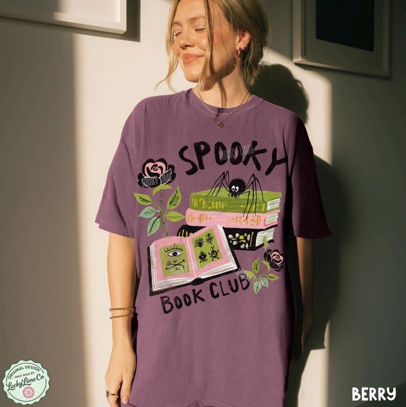 Op de afbeelding: Bessenkleurig T-shirt met een spookachtig boekenclubontwerp. De afbeelding bevat het woord "Spooky", een spin, boeken, rozen en de woorden "Book Club". Het shirt heeft een relaxte pasvorm.