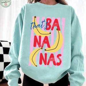 May include: A light blue sweatshirt with the phrase "that's BA NA NAS" in red and blue text, with yellow banana illustrations. The design is set against pink vertical stripes.