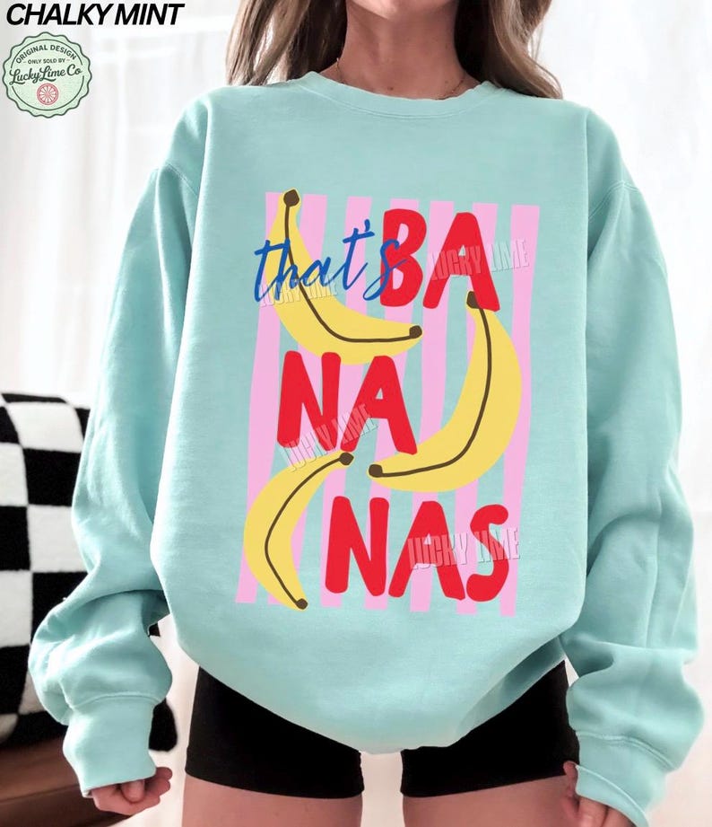 May include: A light blue sweatshirt with the phrase "that's BA NA NAS" in red and blue text, with yellow banana illustrations. The design is set against pink vertical stripes.