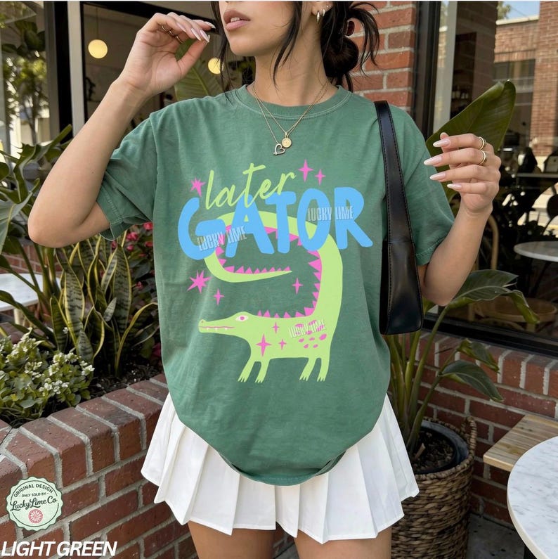 May include: Light green t-shirt with the text "later GATOR" and a cartoon alligator illustration in light green and pink. The shirt is a relaxed fit and has short sleeves. The text "Lucky Lime Co" is at the bottom of the shirt.